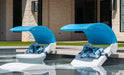 Two white in-pool loungers with blue canopies and patterned pillows partially submerged in a pool with a light stone surround and building in the background.