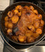 A cast iron Dutch oven filled with a roasted beef brisket and golden potatoes. The meat is browned and glazed, and the potatoes are tender and slightly caramelized. The dish is sitting on a stovetop.