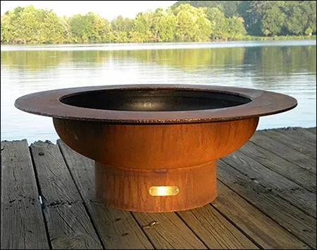 Wood burning fire pit on a wooden deck by a lake. The fire pit is round and made of rusted metal. There is a small light on the bottom of the fire pit.