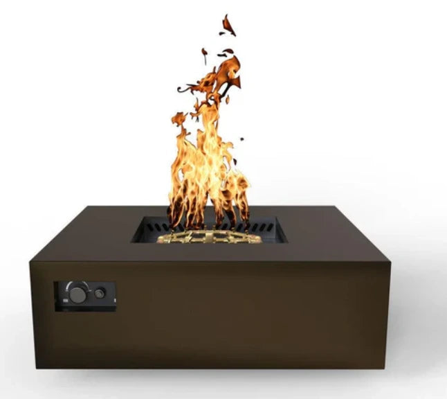 A modern, square gas fire pit table with a bronze finish. Flames dance above the burner, casting a warm glow. Controls are located on the side.