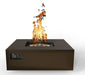 A modern, square gas fire pit table with a bronze finish. Flames dance above the burner, casting a warm glow. Controls are located on the side.