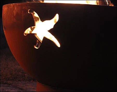 A close-up of a starfish cutout in a round, rusted metal fire pit. Flames are visible through the cutout.