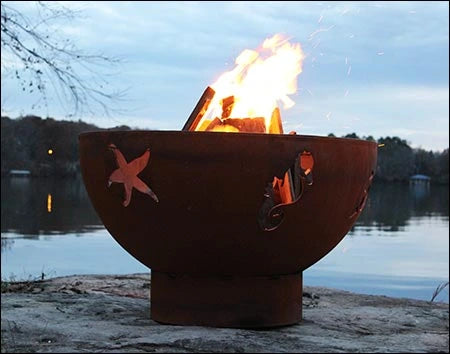 A round, rusted metal fire pit with flames burning inside. The fire pit has a seahorse and starfish design cut into the metal. It is placed on a rock near a body of water at sunset.