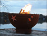 A round, rusted metal fire pit with flames burning inside. The fire pit has a seahorse and starfish design cut into the metal. It is placed on a rock near a body of water at sunset.