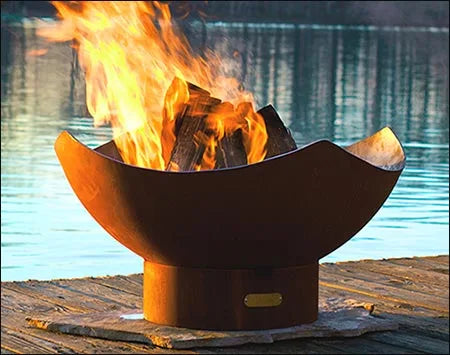  A large, round fire pit made of rusted metal is sitting on a wooden dock. The fire pit is filled with burning wood, and flames are leaping high into the air. A body of water is visible in the background.