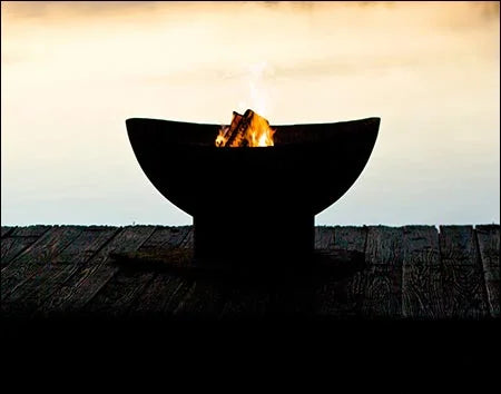 A round, black fire pit with flames burning inside. The fire pit is placed on a wooden dock near a body of water at sunset.