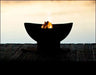 A round, black fire pit with flames burning inside. The fire pit is placed on a wooden dock near a body of water at sunset.