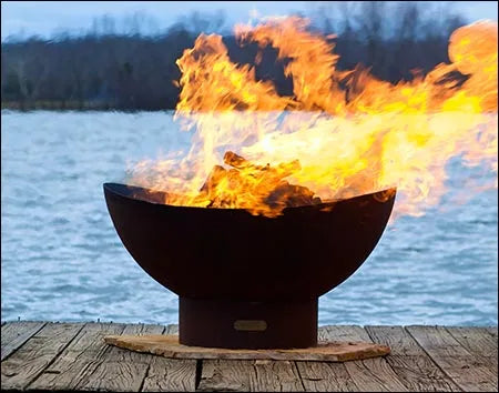 A round, rusted metal fire pit with flames burning inside. The fire pit is placed on a wooden dock near a body of water. The sky is dark and the water is reflecting the flames.