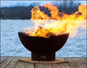 A round, rusted metal fire pit with flames burning inside. The fire pit is placed on a wooden dock near a body of water. The sky is dark and the water is reflecting the flames.