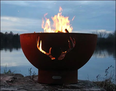 Wood burning fire pit with a deer antler cutout design. The fire pit is round and made of rusted metal. Flames are burning brightly inside, and the fire pit is placed on a rock near a lake. The sky is dark with a hint of orange from the sunset.