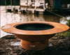 A large, round, rust-colored fire pit with a raised outer rim. The fire pit is placed on a rock near a body of water. There are boats docked in the background.