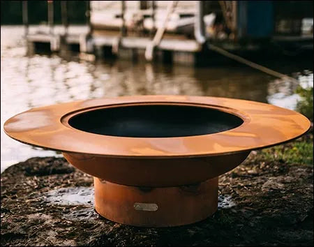 A large, round, rust-colored fire pit with a raised outer rim. The fire pit is placed on a rock near a body of water. There are boats docked in the background.