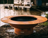 A large, round, rust-colored fire pit with a raised outer rim. The fire pit is placed on a rock near a body of water. There are boats docked in the background.