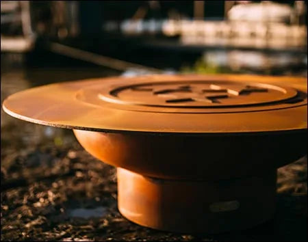 A close-up of a round, rust-colored wood-burning fire pit with a circular design in the center. The fire pit is placed on a rock near a body of water.