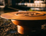 A close-up of a round, rust-colored wood-burning fire pit with a circular design in the center. The fire pit is placed on a rock near a body of water.