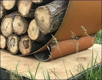 Close-up of a circular steel firewood rack with firewood stacked inside. The rack is resting on a wooden platform and has a rusted finish.