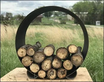 Carbon steel circular firewood rack with firewood stacked inside. The rack is standing on a wooden platform in a grassy area.