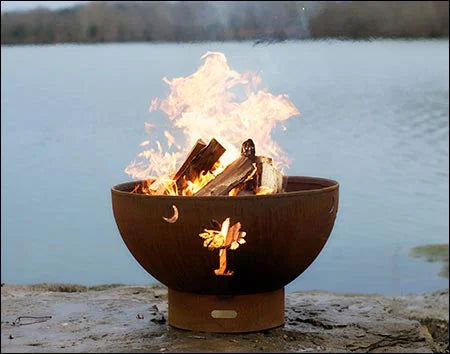 A round, rust-colored wood-burning fire pit with a palm tree cutout. Flames are burning inside the pit, and it is placed on a rock near a body of water.