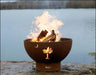 A round, rust-colored wood-burning fire pit with a palm tree cutout. Flames are burning inside the pit, and it is placed on a rock near a body of water.