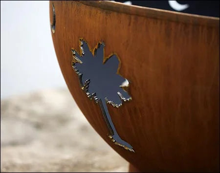 A close-up of a palm tree cutout in a rust-colored fire pit. The cutout is filled with shadow, creating a contrast against the warm metal.