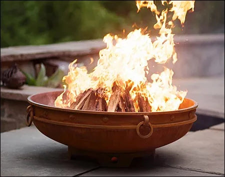 A round, rusted metal fire pit with a large, roaring fire burning inside. The fire pit is placed on a patio near a body of water. There are tall grasses and a stone wall in the background.