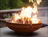 A round, rusted metal fire pit with a large, roaring fire burning inside. The fire pit is placed on a patio near a body of water. There are tall grasses and a stone wall in the background.