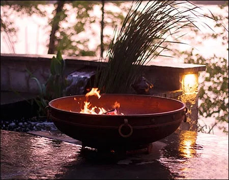 A round fire pit with flames burning inside. The fire pit is placed on a patio near a body of water. There are tall grasses and a stone wall in the background.