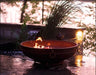 A round fire pit with flames burning inside. The fire pit is placed on a patio near a body of water. There are tall grasses and a stone wall in the background.