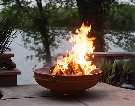 A wood-burning fire pit with a large, roaring flame. The fire pit is made of rusted metal and has handles on the sides.