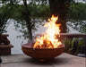 A wood-burning fire pit with a large, roaring flame. The fire pit is made of rusted metal and has handles on the sides.