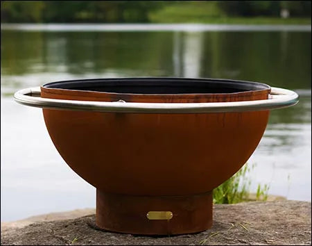 A round, rust-colored wood burning fire pit with a stainless steel handle. It is sitting on a rock near a body of water.