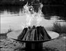 A large, round stainless steel wood burning fire pit with a roaring fire in the center. The fire pit is placed on a rocky shoreline near a lake with trees in the background. The image is in black and white.