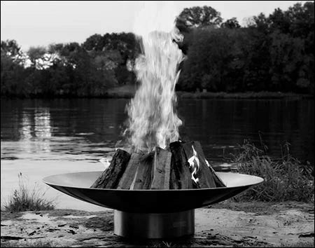 A large, round stainless steel wood burning fire pit with a roaring fire in the center. The fire pit is placed on a rocky shoreline near a lake with trees in the background. The image is in black and white.