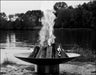 A large, round stainless steel wood burning fire pit with a roaring fire in the center. The fire pit is placed on a rocky shoreline near a lake with trees in the background. The image is in black and white.