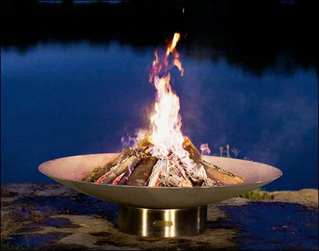 A large, round wood burning fire pit with a roaring fire in the center. The fire pit is placed on a stone patio overlooking a body of water with a dark blue sky in the background.