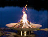 A large, round wood burning fire pit with a roaring fire in the center. The fire pit is placed on a stone patio overlooking a body of water with a dark blue sky in the background.