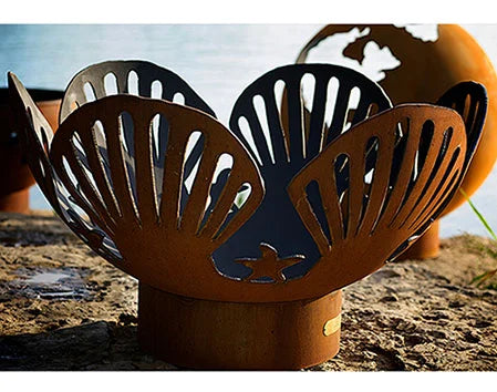 A close-up of a seashell-shaped fire pit made of rusted metal. The fire pit has intricate cutouts and a starfish design.