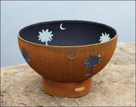 A round, rust-colored fire pit with palm tree and crescent moon cutouts. The cutouts are filled with white light, creating a contrast against the dark background.