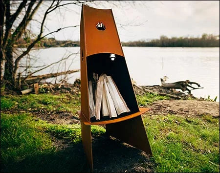  A tall, conical fire pit with firewood inside. The fire pit is surrounded by green grass and is near a body of water.