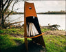  A tall, conical fire pit with firewood inside. The fire pit is surrounded by green grass and is near a body of water.