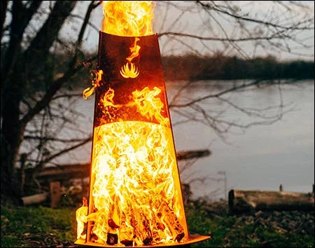 A tall, conical wood-burning fire pit with flames burning inside. The fire pit is surrounded by green grass and is near a body of water. There is a deer logo on the side of the fire pit.