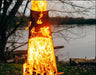 A tall, conical wood-burning fire pit with flames burning inside. The fire pit is surrounded by green grass and is near a body of water. There is a deer logo on the side of the fire pit.