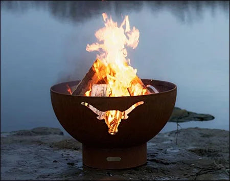  A wood-burning fire pit with a longhorn cutout design, placed on a rocky shoreline near a lake with a misty background. The fire is burning brightly.