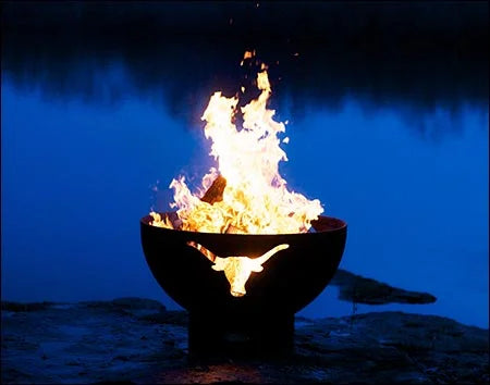 A wood-burning fire pit with a longhorn cutout design, placed on a rocky shoreline near a lake. The fire is burning brightly, with flames reaching high into the night sky.