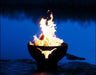 A wood-burning fire pit with a longhorn cutout design, placed on a rocky shoreline near a lake. The fire is burning brightly, with flames reaching high into the night sky.