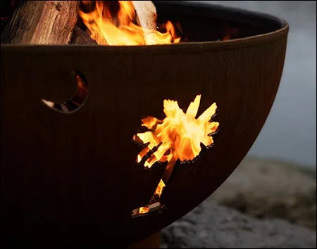 A close-up of a palm tree cutout in a rust-colored fire pit. Flames are burning through the cutout, casting a warm glow.