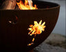 A close-up of a palm tree cutout in a rust-colored fire pit. Flames are burning through the cutout, casting a warm glow.