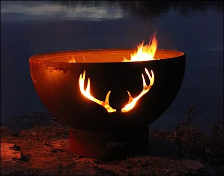 Wood burning fire pit with a deer antler cutout design. The fire pit is round and made of rusted metal. Flames are burning brightly inside, casting a warm glow on the deer antler silhouette. The fire pit is placed on a rock near a lake at night.