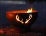 Wood burning fire pit with a deer antler cutout design. The fire pit is round and made of rusted metal. Flames are burning brightly inside, casting a warm glow on the deer antler silhouette. The fire pit is placed on a rock near a lake at night.