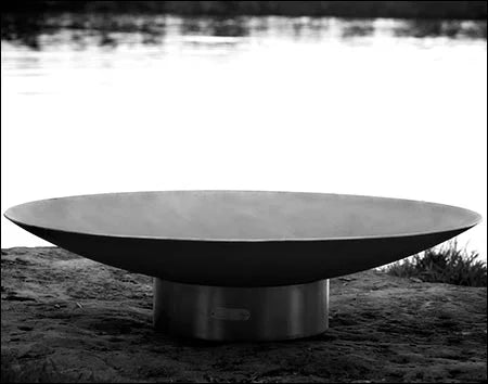 A large, round steel wood burning fire pit with a smooth, reflective surface. The fire pit is placed on a rocky shoreline near a lake.